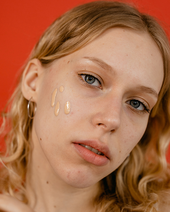 Mladá žena má na tvári tri kvapky tekutého minerálneho make-upu a stojí pred červeným pozadím.